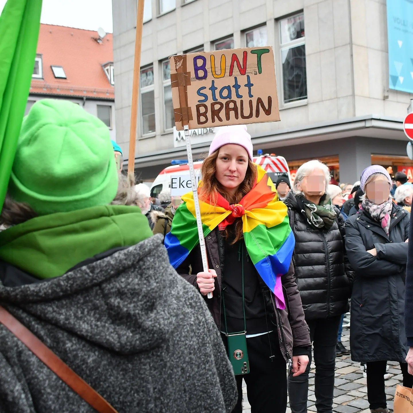 Rebecca Lenhard steht auf einer Demo mit Regenbogenflagge um die Schultern und Schild mit "BUNT statt BRAUN" Poster in der Hand.